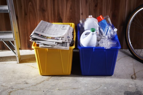 In a garage or storage area, there are two large plastic recycling bins placed side by side against a wooden wall. The left bin, which is yellow, contains a pile of assorted newspapers and magazines with visible text and images on the pages. The right bin, blue in colour, holds clear plastic bottles, some empty and partially crushed, along with a few plastic containers and a white plastic jug. The concrete floor beneath the bins shows slight wear and some minor cracks. In the background, part of a metal shelving unit is visible on the left, and the edge of a bicycle wheel can be seen on the right, indicating that this space is used for storage and disposal of various household waste materials. The scene is illuminated by natural or ambient lighting, highlighting the textures of the plastic, paper, and wooden surroundings, and reflects typical rubbish management practices for local households or small businesses, such as those served by companies like Office Clearance Honor Oak in South London postcode area.