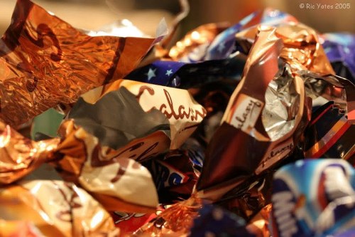 A close-up view of a pile of crumpled plastic wrappers and foil packaging, featuring various metallic, glossy, and matte finishes in shades of copper, silver, and multicolored accents. The wrappers appear to be discarded confectionery or snack packaging, with some displaying partially visible text and logos. The background suggests an indoor setting or a collection of rubbish awaiting removal, with the wrappers overlapping each other in a disorderly manner. The lighting highlights the reflective surfaces and textured creases of the packaging materials. This scene exemplifies waste related to packaged snack foods, aligning with rubbish removal services offered by Office Clearance Honor Oak, serving areas around London postcodes near Honor Oak. The image emphasizes the importance of proper disposal and the type of waste managed by professional rubbish clearance services aimed at maintaining cleanliness and waste management in local communities.