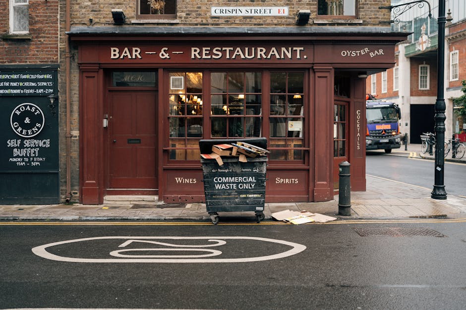 In front of a brick-built restaurant with a reddish-brown facade and large glass windows, a black wheeled commercial waste bin marked 'COMMERCIAL WASTE ONLY' is positioned on the pavement, overflowing