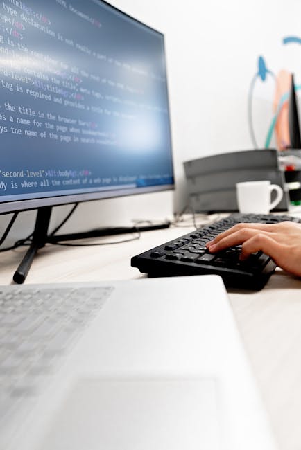 In a modern office environment, a person with short hair is seated at a desk, facing a large computer monitor displaying lines of colorful computer code on a dark background. The workspace is well-lit