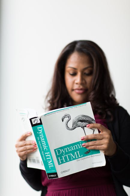 A woman with medium-length dark hair and medium skin tone is standing indoors against a plain white background, holding a thick book titled 'Dynamic HTML' with a turquoise label and an image of a flam